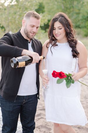 Man pouring champagne for woman holding red roses during a romantic beach engagement.