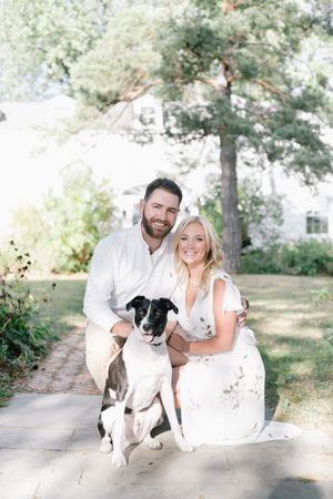 Smiling engaged couple posing outdoors with their black and white dog on a garden path.
