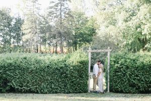 Engaged couple kissing under a rustic wooden arch surrounded by lush green hedges.