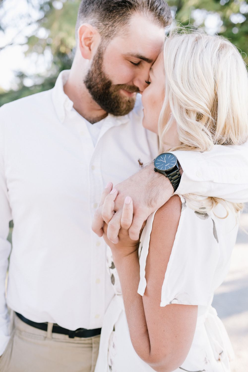 Bearded man in white shirt kissing blonde woman while holding hands outdoors in soft light.