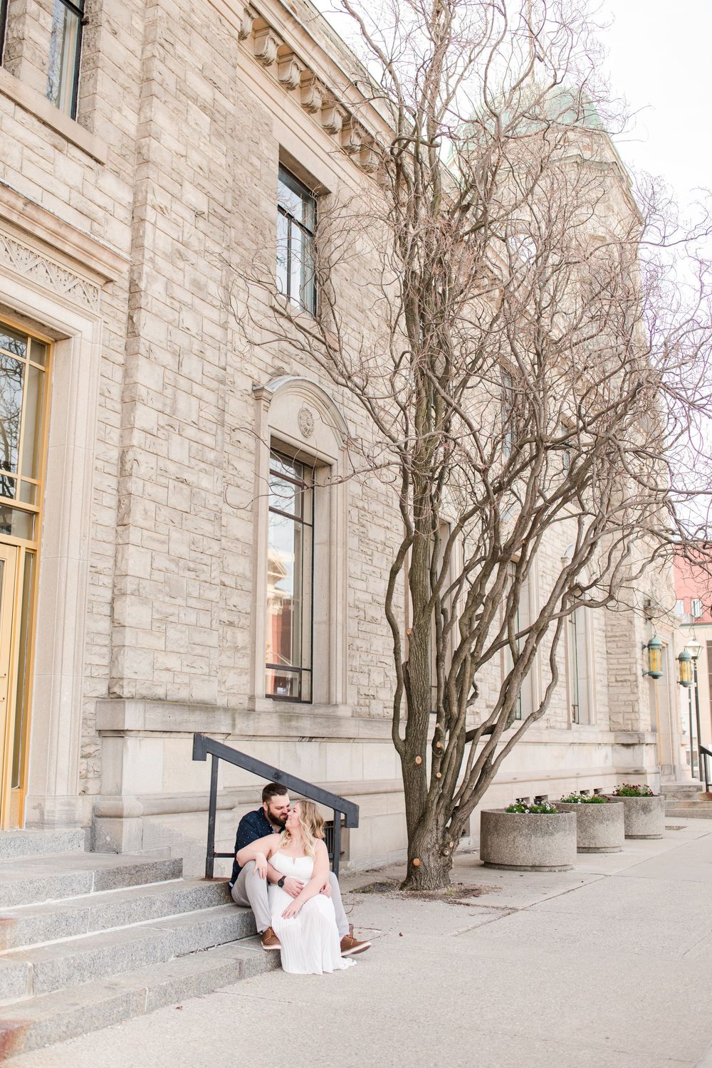 Couple sitting on stone steps embracing outside a historic limestone building with a bare tree in spring.