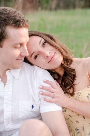Woman resting her head on man's shoulder showing engagement ring in a sunny field.