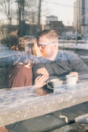Couple kissing over coffee cups at a cafe table, photographed through a sunlit window.