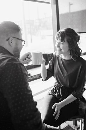 Black and white photo of a couple sharing coffee and smiling at each other by a window.