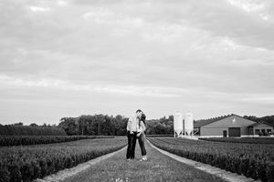 Couple sharing a romantic kiss on a farm pathway surrounded by lush green fields in black and white.