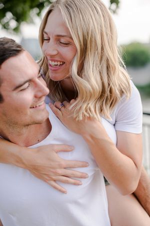 Happy couple laughs together, woman with blonde hair hugs man from behind outdoors.