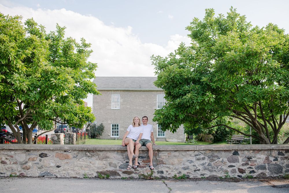 Couple sits on a stone wall surrounded by lush green trees and a historic stone building.