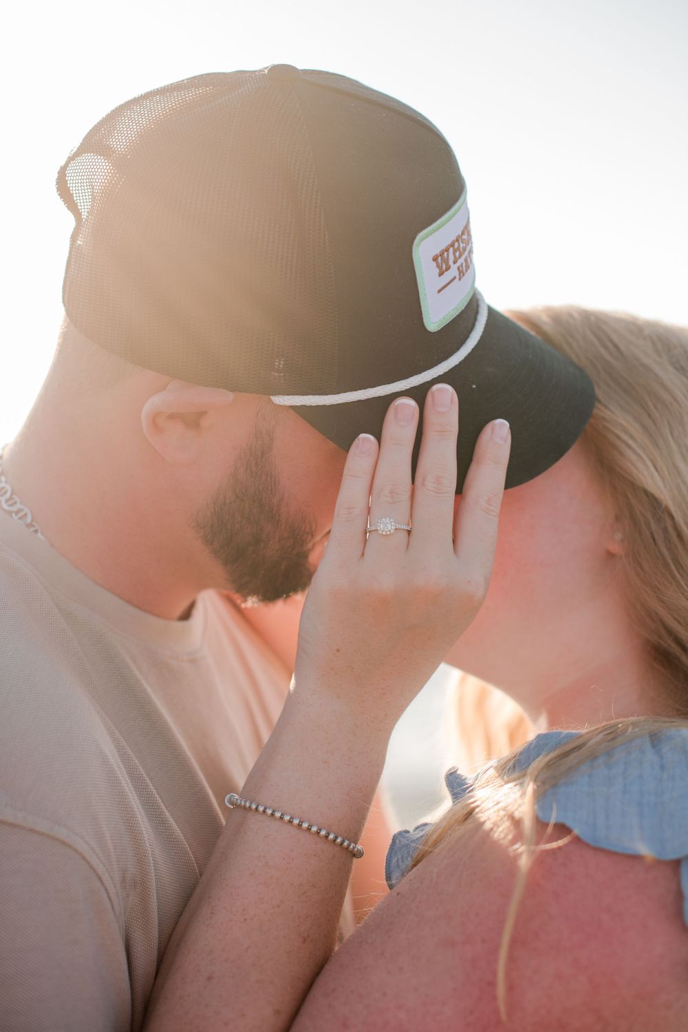 Couple kissing at sunset while woman displays sparkling engagement ring toward camera.