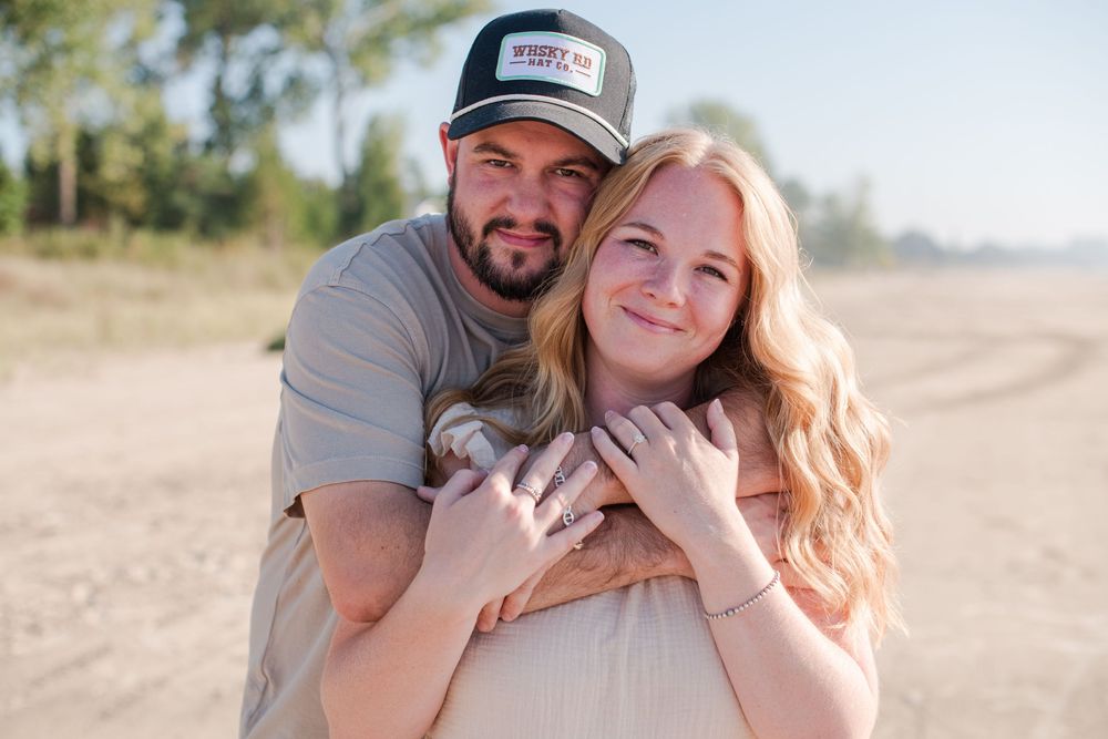 Man hugging woman from behind as they smile together on a sandy outdoor path.