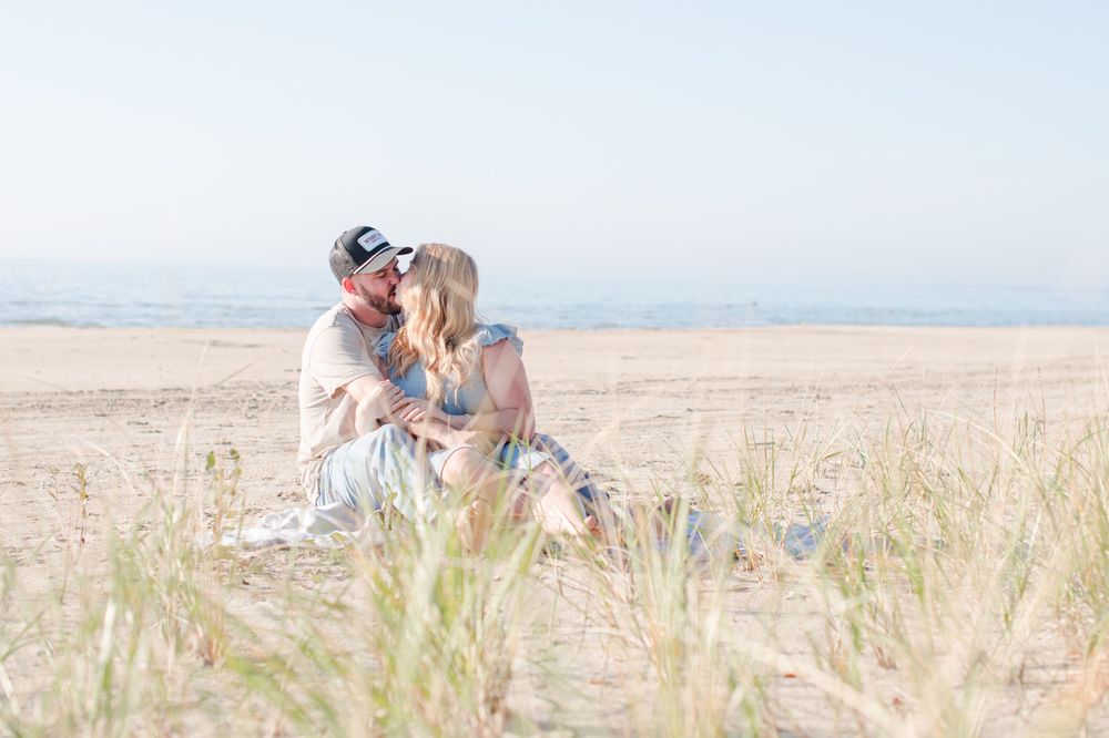 Couple sitting together kissing on sandy beach dunes with tall grass in the foreground.