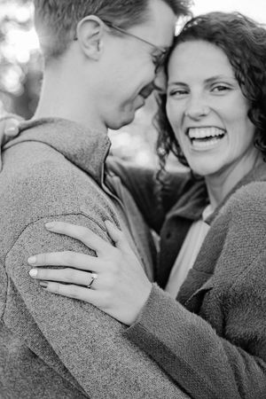 Black and white close-up of laughing woman embracing man, engagement ring visible on her hand.