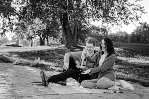 Black and white photo of couple sitting on blanket under trees sharing a bottle of champagne.