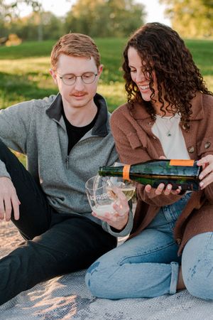 Couple sitting on blanket outdoors as woman pours champagne into glass during engagement celebration.