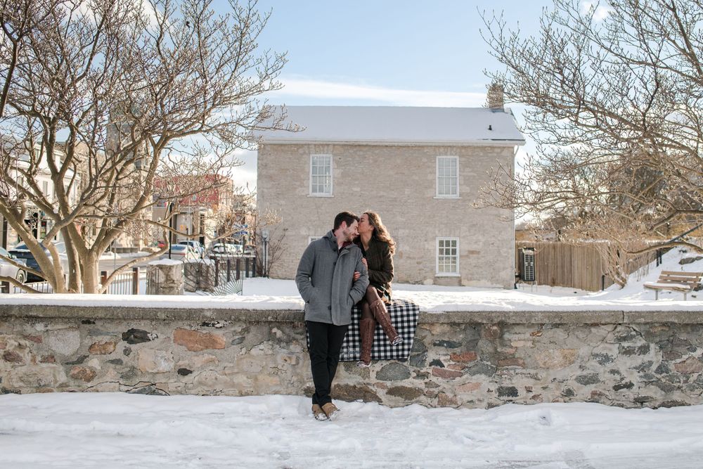 Couple sharing a romantic moment on a snowy stone wall outside a historic building in winter.