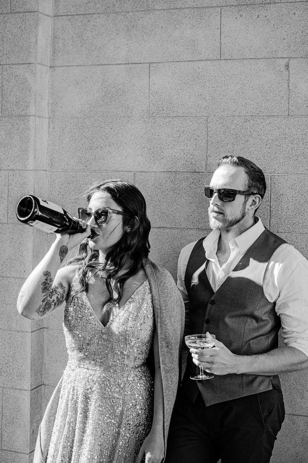 Black and white photo of bride drinking from bottle and groom holding glass in sunglasses.