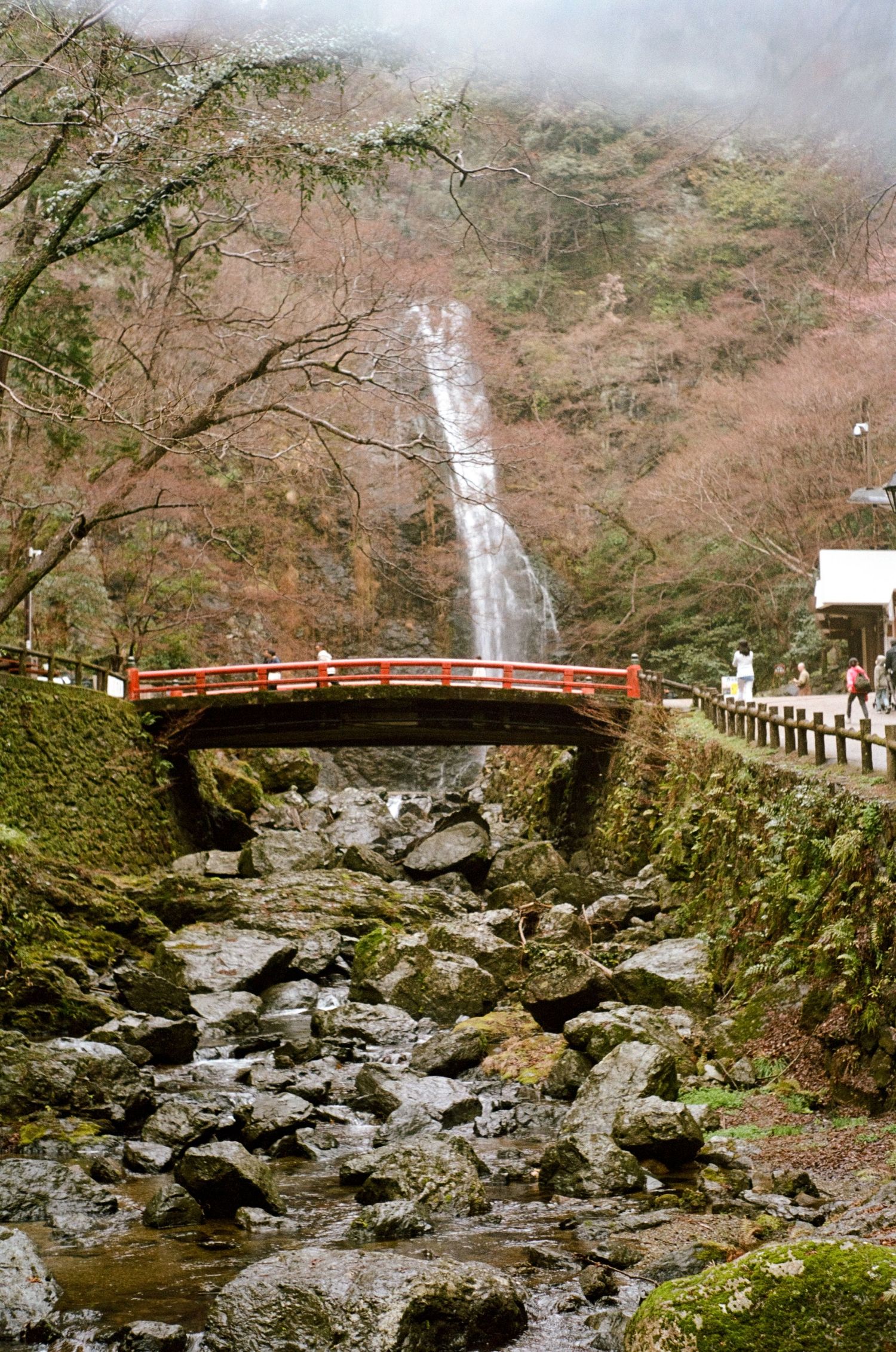 Red bridge spans rocky stream beneath tall waterfall in misty Japanese forest.