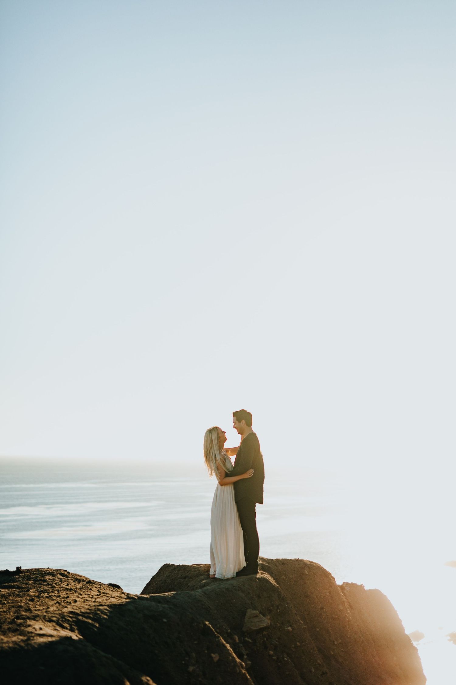 Couple embracing on rocky cliffs at sunset with ocean horizon backdrop during engagement photo session.