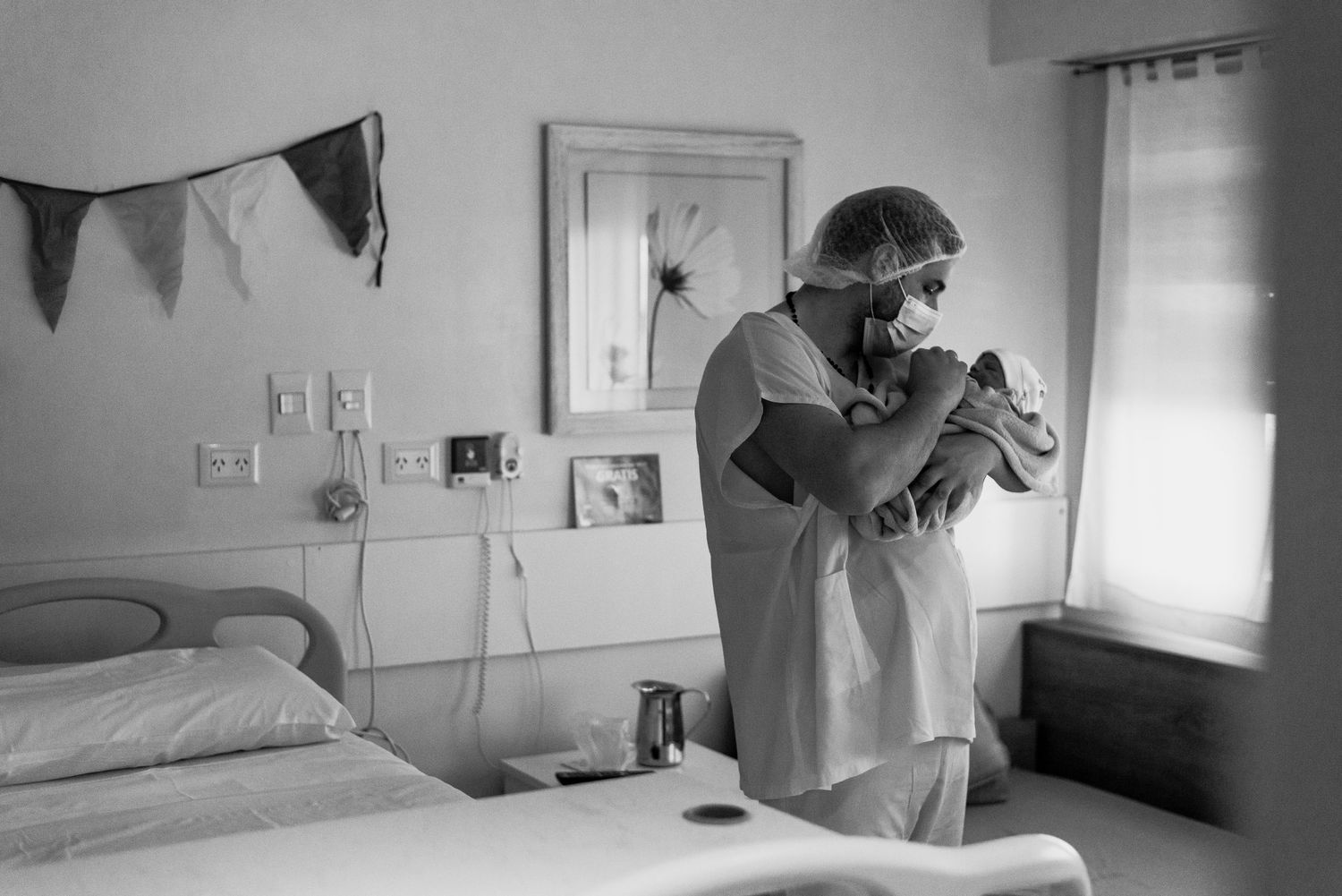 Padre con ropa de quirófano en habitación de sanatorio sosteniendo y mirando a sus bebé recién nacido