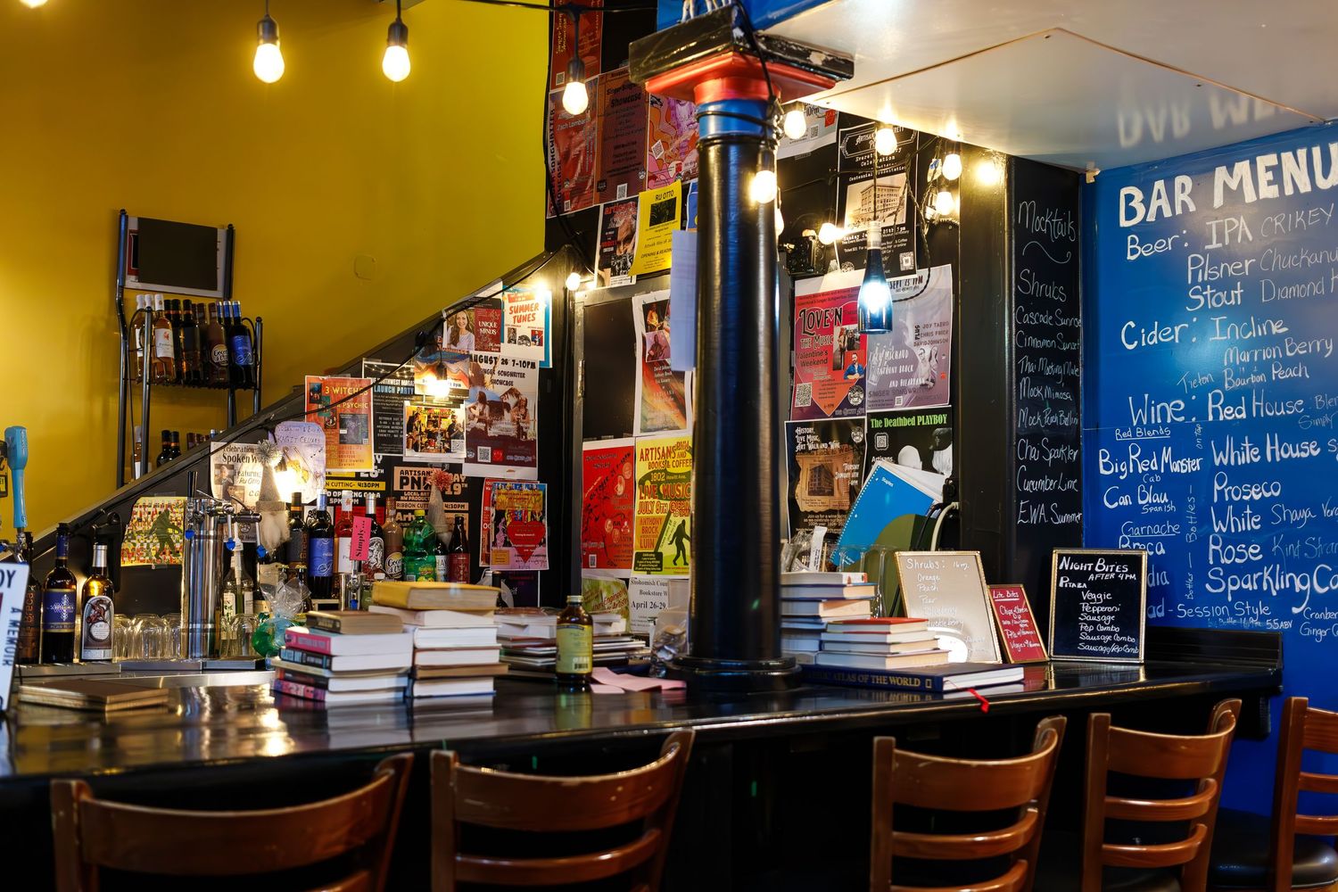 Cozy bookstore bar with wooden chairs and chalkboard menu displaying drink options.