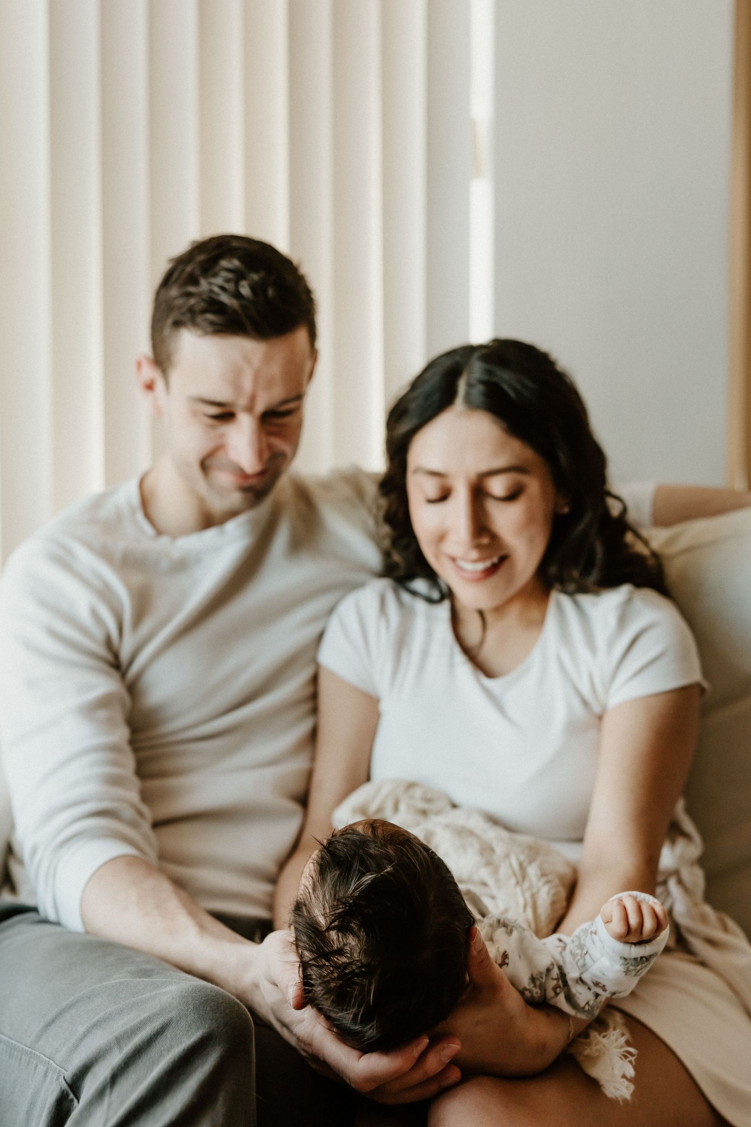 A couple in white tops share an intimate moment with a baby on a couch in a bright minimalist setting.