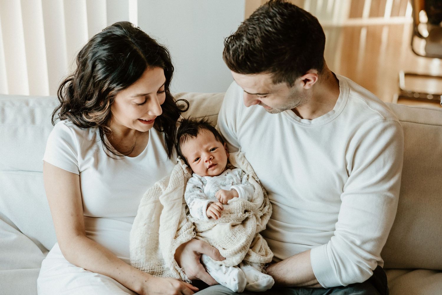 A family sits together on a light-colored couch, lovingly gazing at their laughing baby in white clothing.