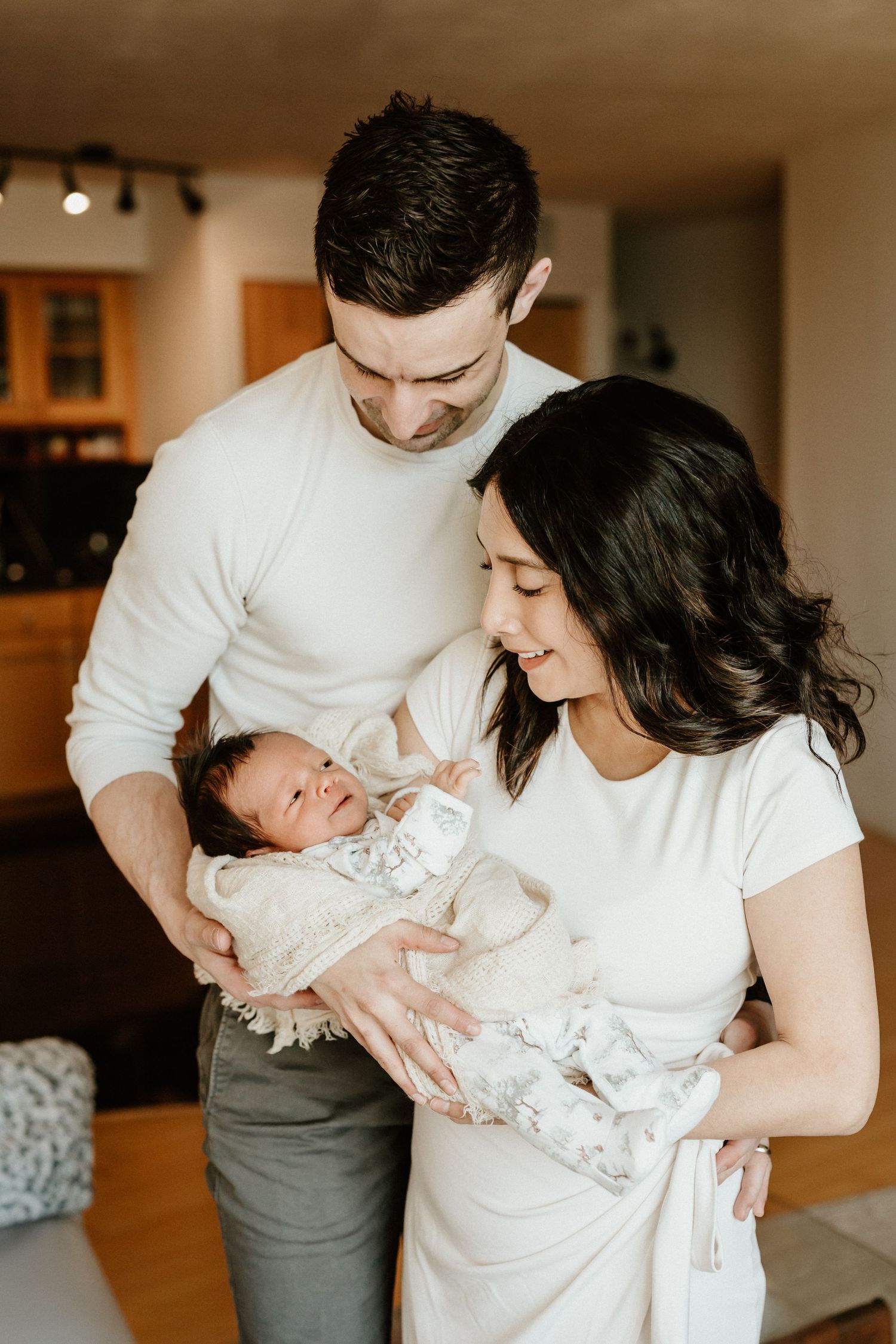 A couple in white shirts holding their newborn baby in a cozy home setting with warm lighting.