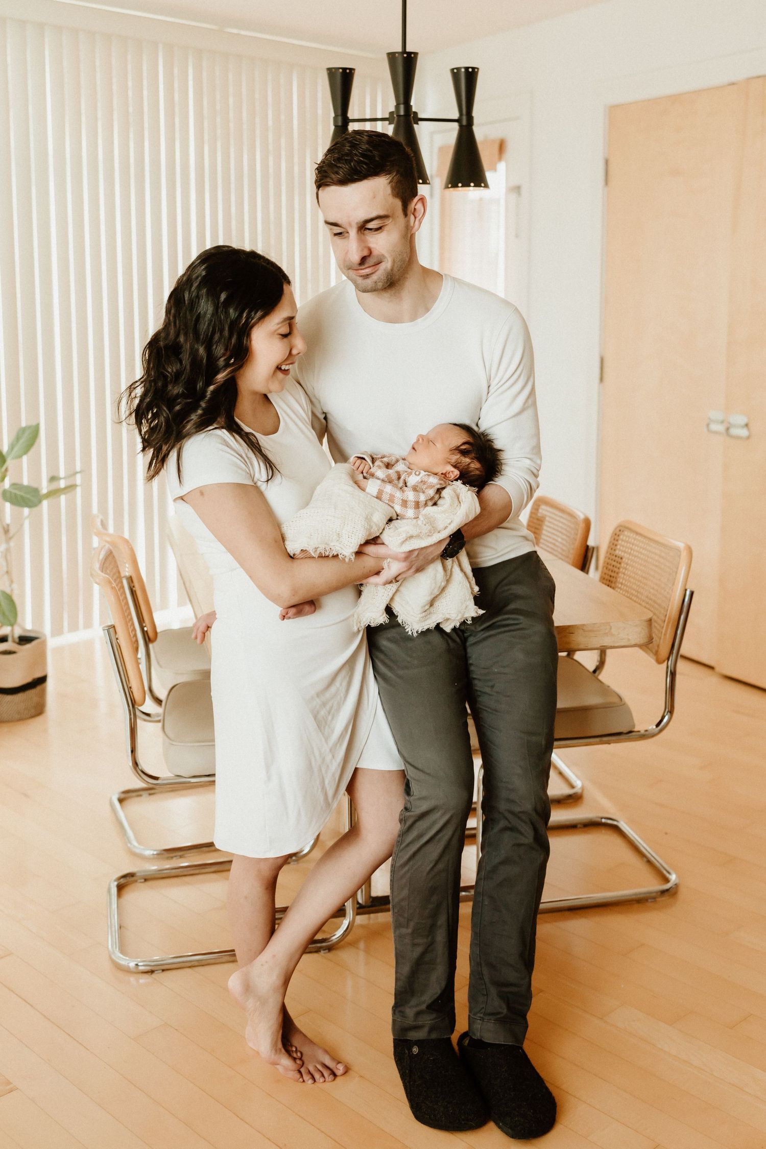 A young couple in casual attire stands together in their modern dining room holding a newborn baby.