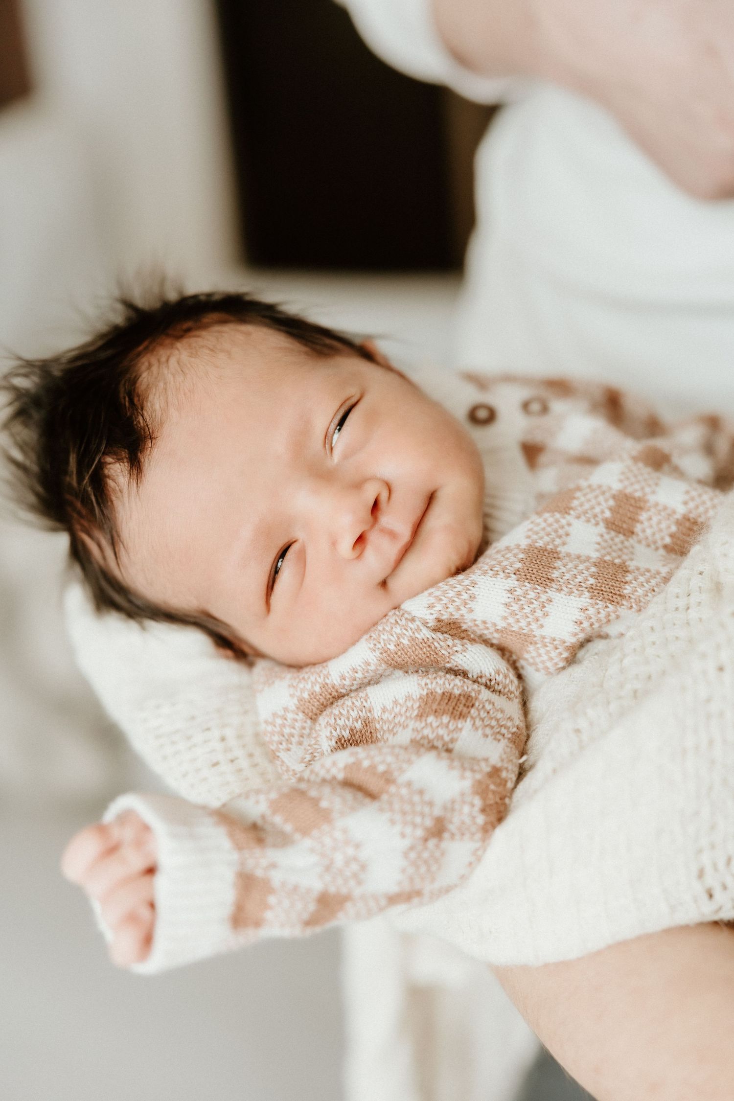 A newborn baby with dark hair wearing a checkered sweater smiles softly while being held.