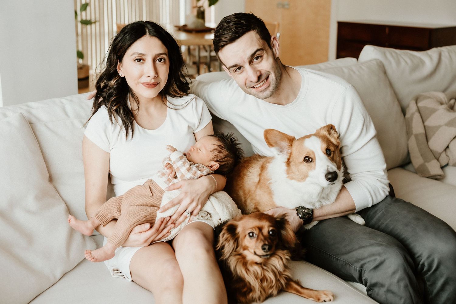 A couple sits on a couch with their infant baby and two dogs, including a corgi and a small brown dog.
