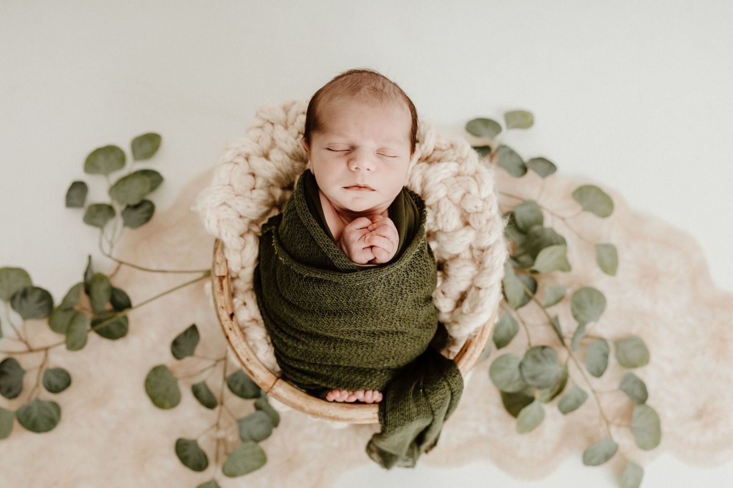 Newborn baby sleeping in a woven basket surrounded by eucalyptus leaves and cozy knits.