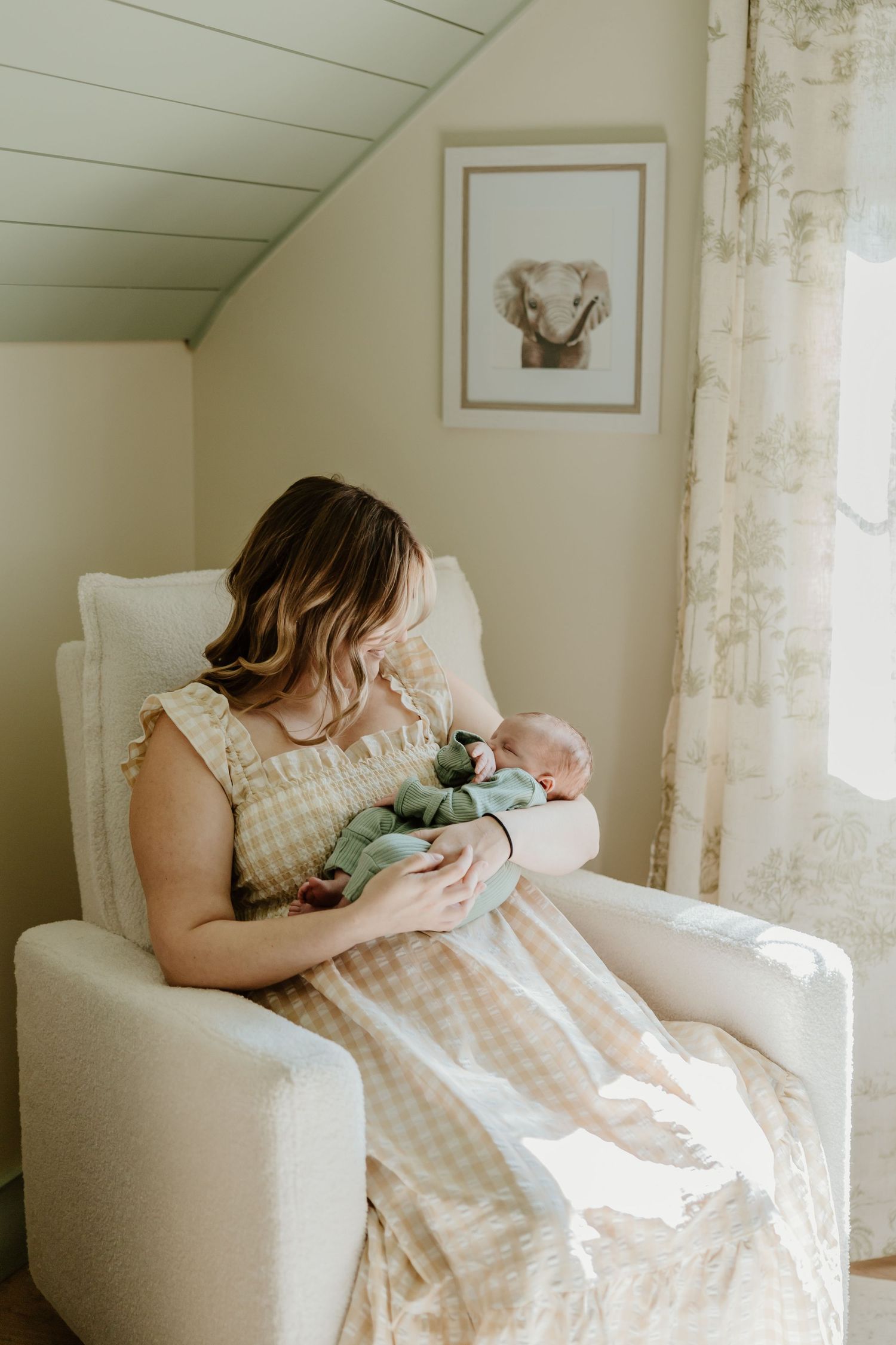 A tender nursery moment in a white armchair beneath framed wall art in a softly lit room.