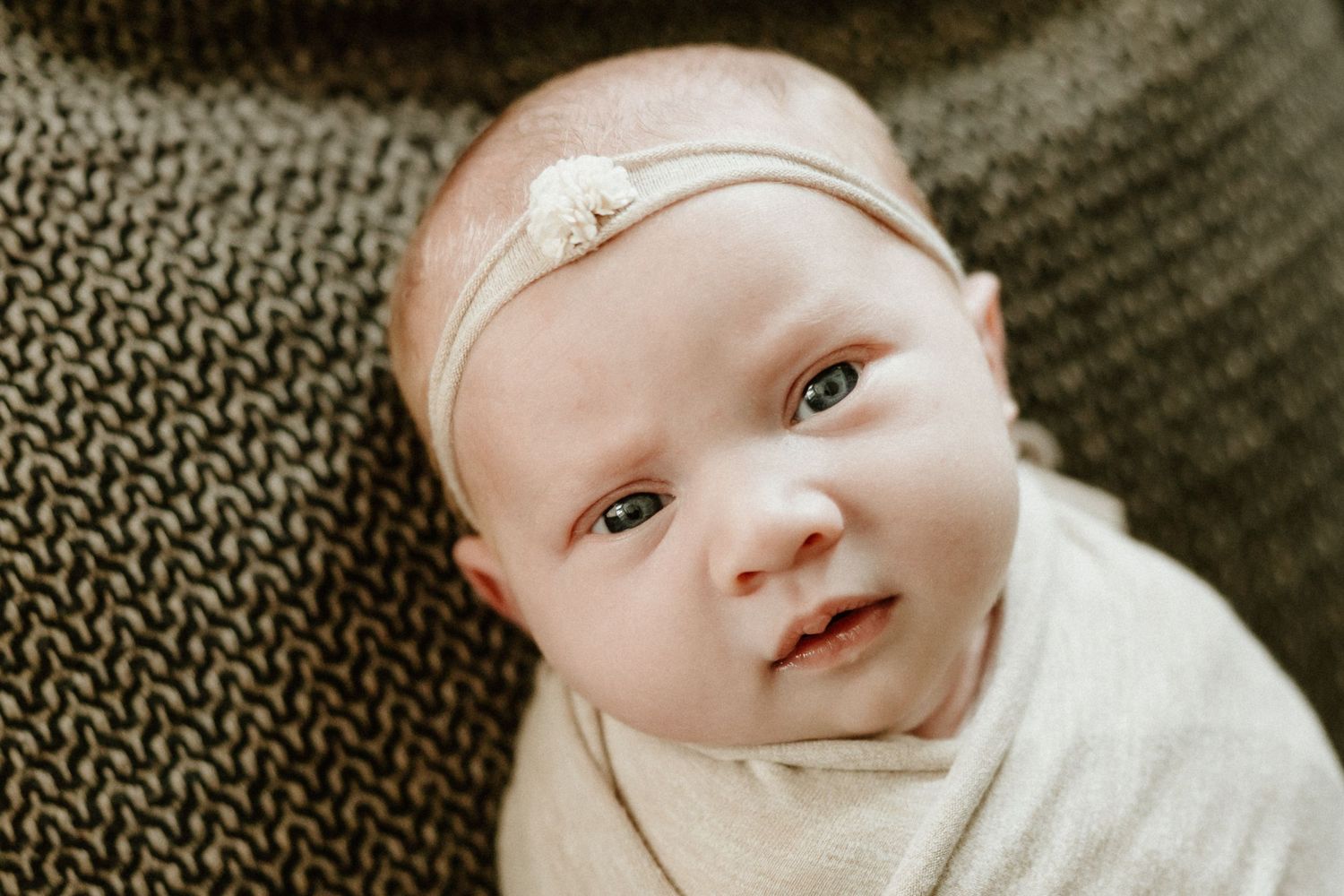 Close-up portrait of baby wearing white with delicate headband against textured gray background.