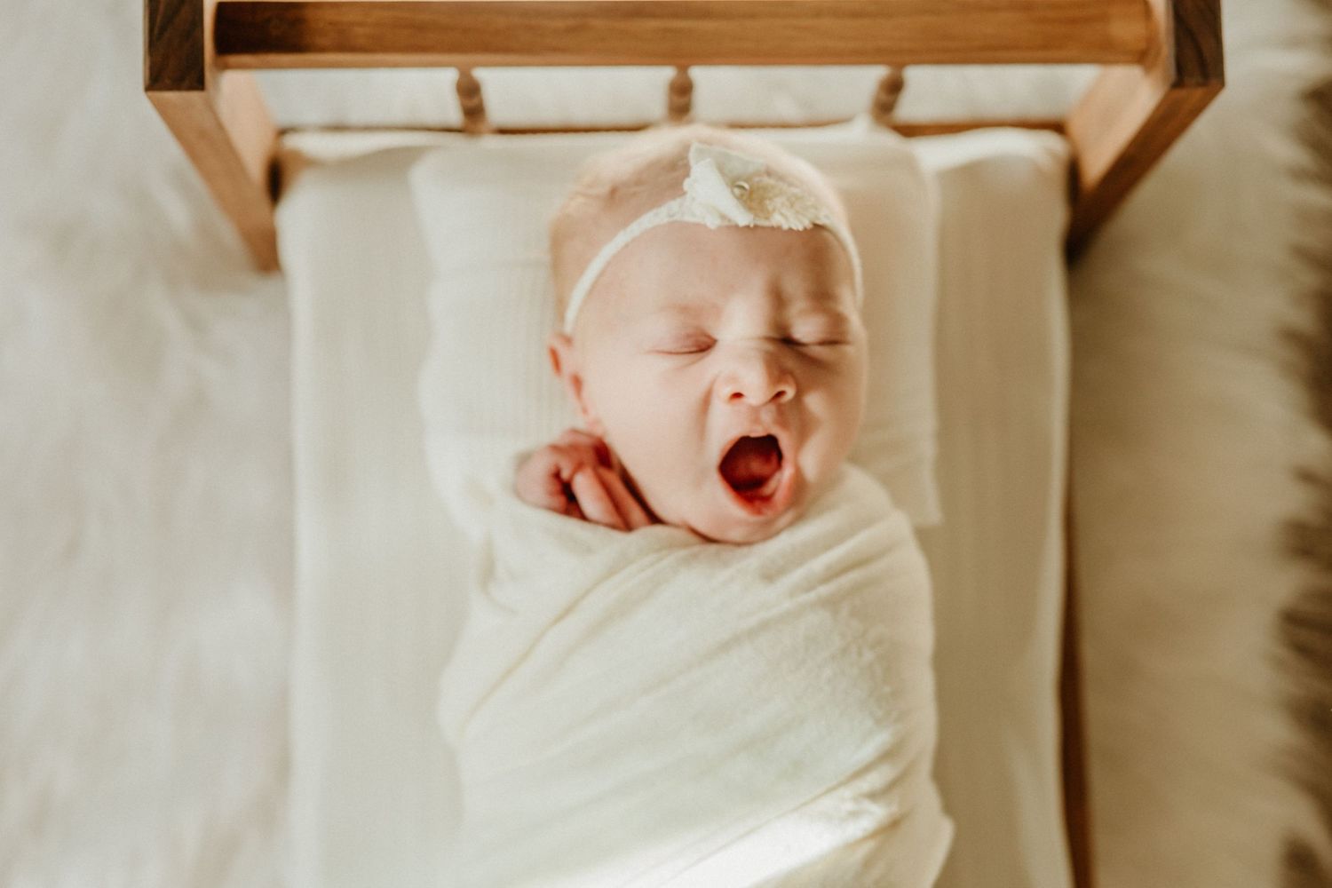 Newborn baby yawning while swaddled in white fabric against a soft backdrop.