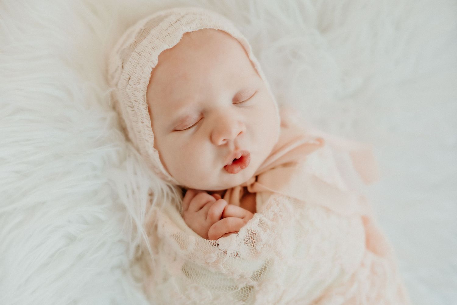 Peaceful sleeping newborn wrapped in delicate cream lace on white fur.