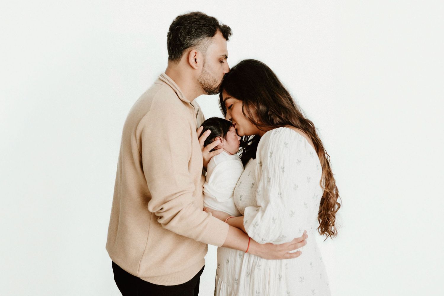 Loving parents embrace while holding their newborn baby against white background.