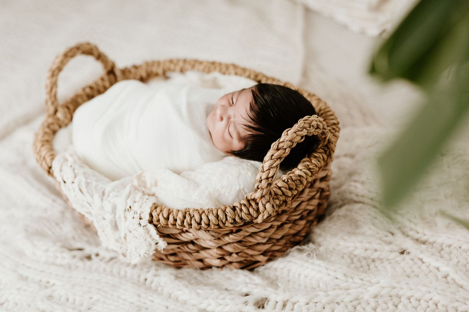 Sleeping newborn wrapped in white blanket rests in woven basket.