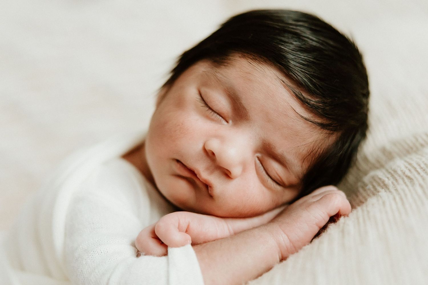 Close-up of sleeping newborn with dark hair wearing white outfit.