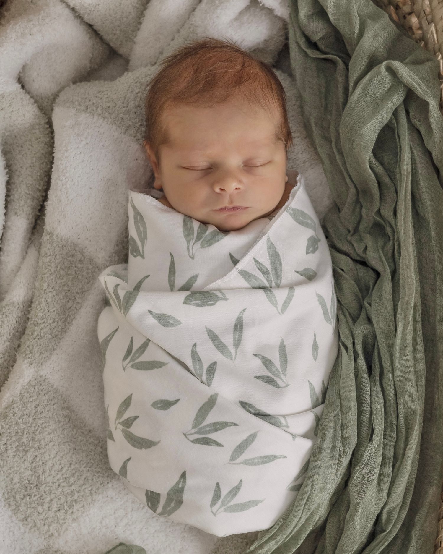 Newborn baby sleeping under a white and sage green leaf-patterned swaddle blanket.