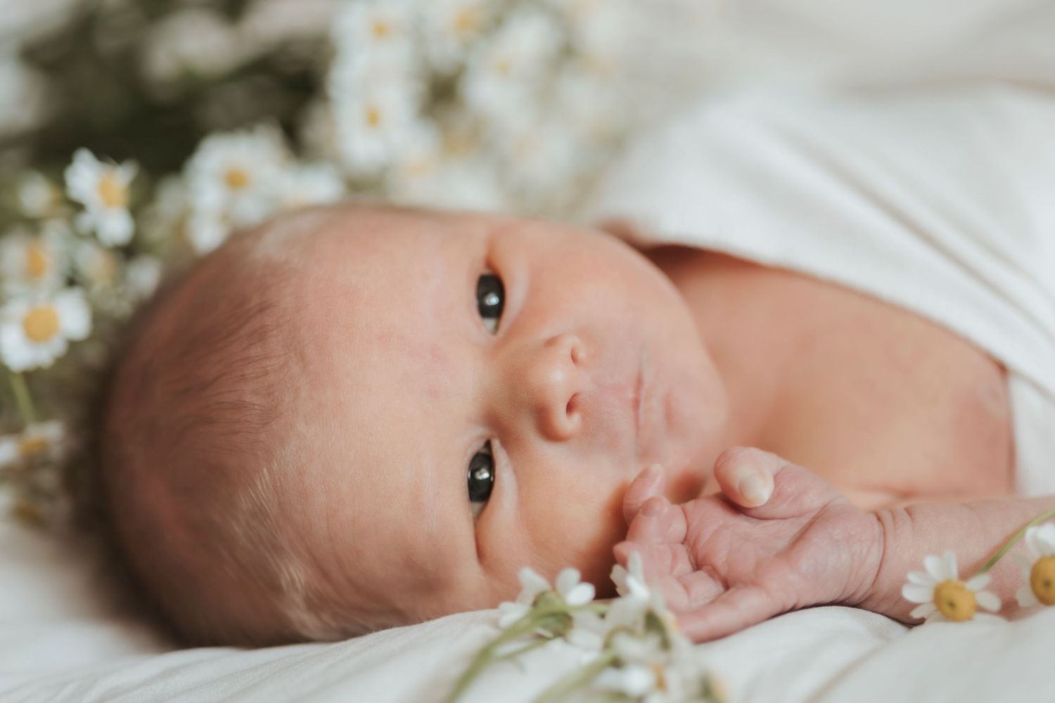 A newborn lying on white fabric with small daisies, gazing at the camera.