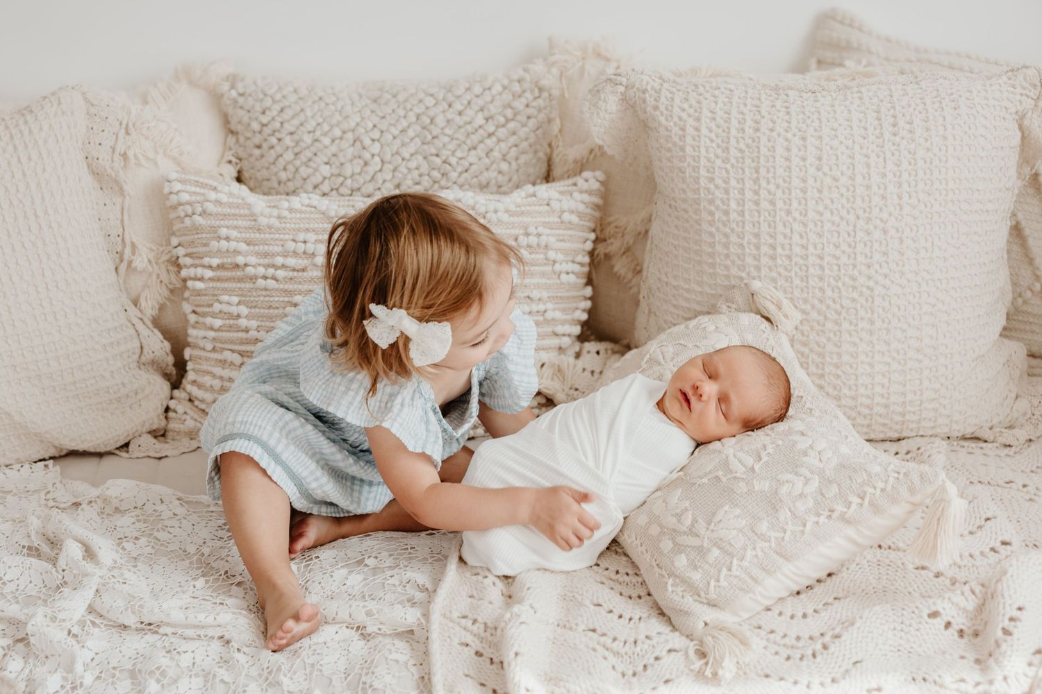Two siblings share a tender moment on a white bed with textured pillows.