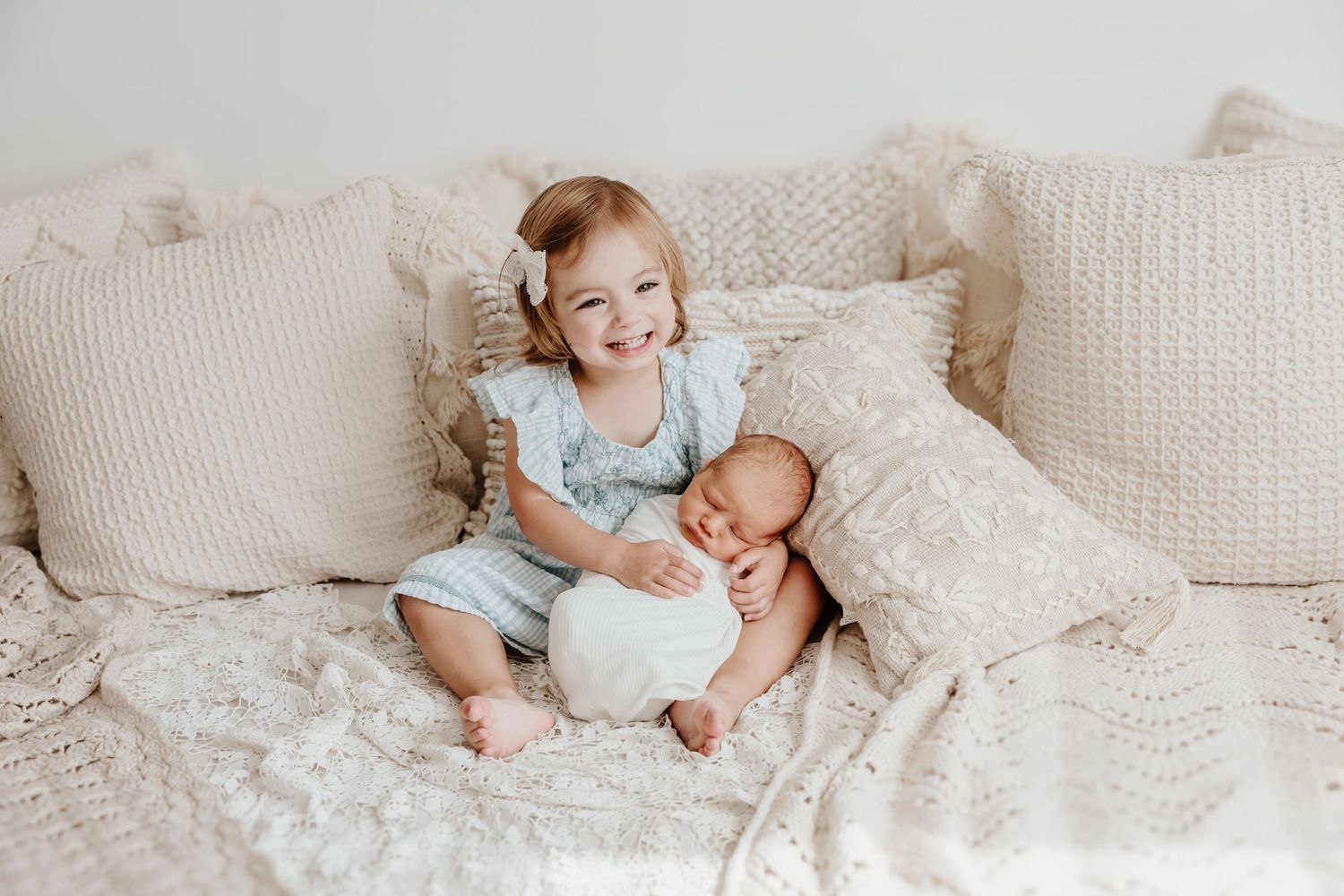 Two children sit together on a cozy white bed with decorative pillows.