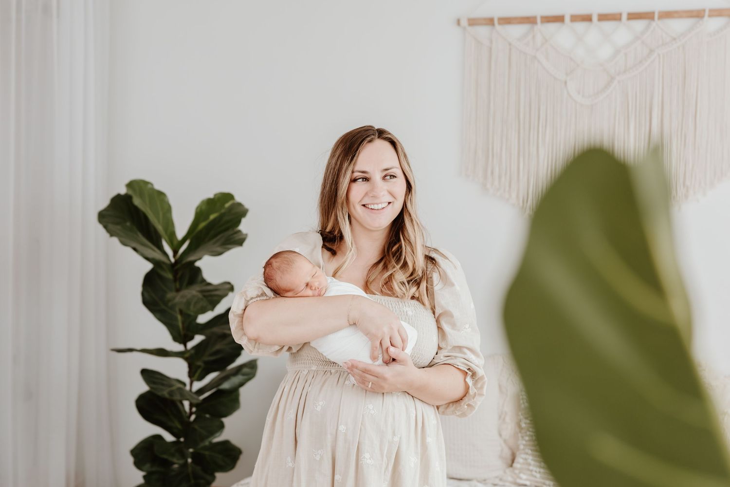 A woman in a cream dress holds her newborn baby next to a fiddle leaf fig plant.