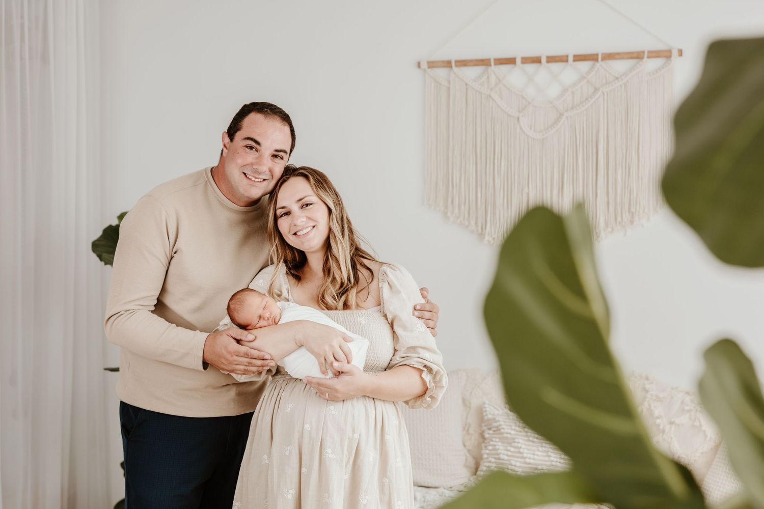 A family poses together with their newborn baby in front of a macrame wall hanging.