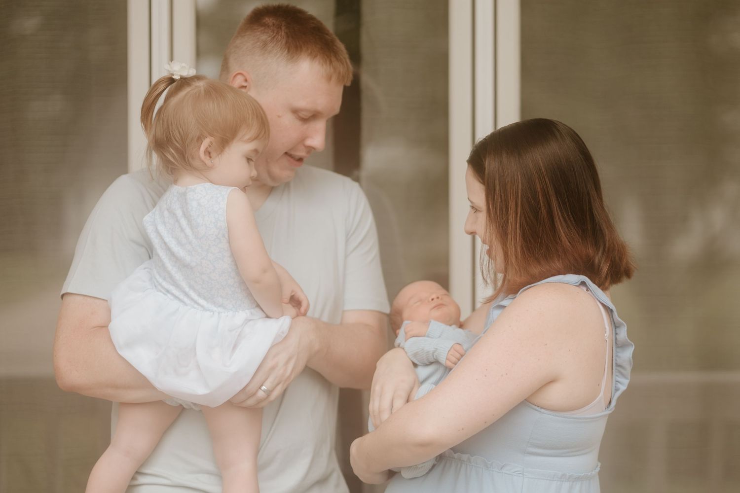 A young couple in casual clothing hold their two small babies while standing together indoors.
