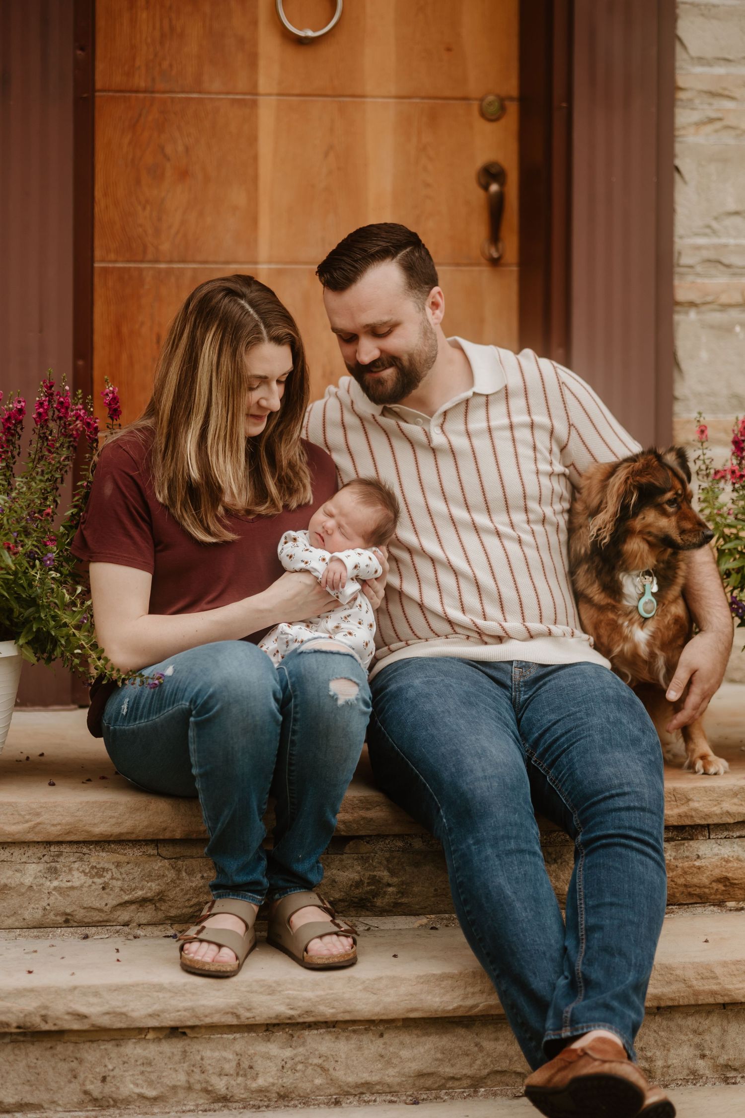 Young family and dog sitting together on stone steps in front of wooden door with flowers.