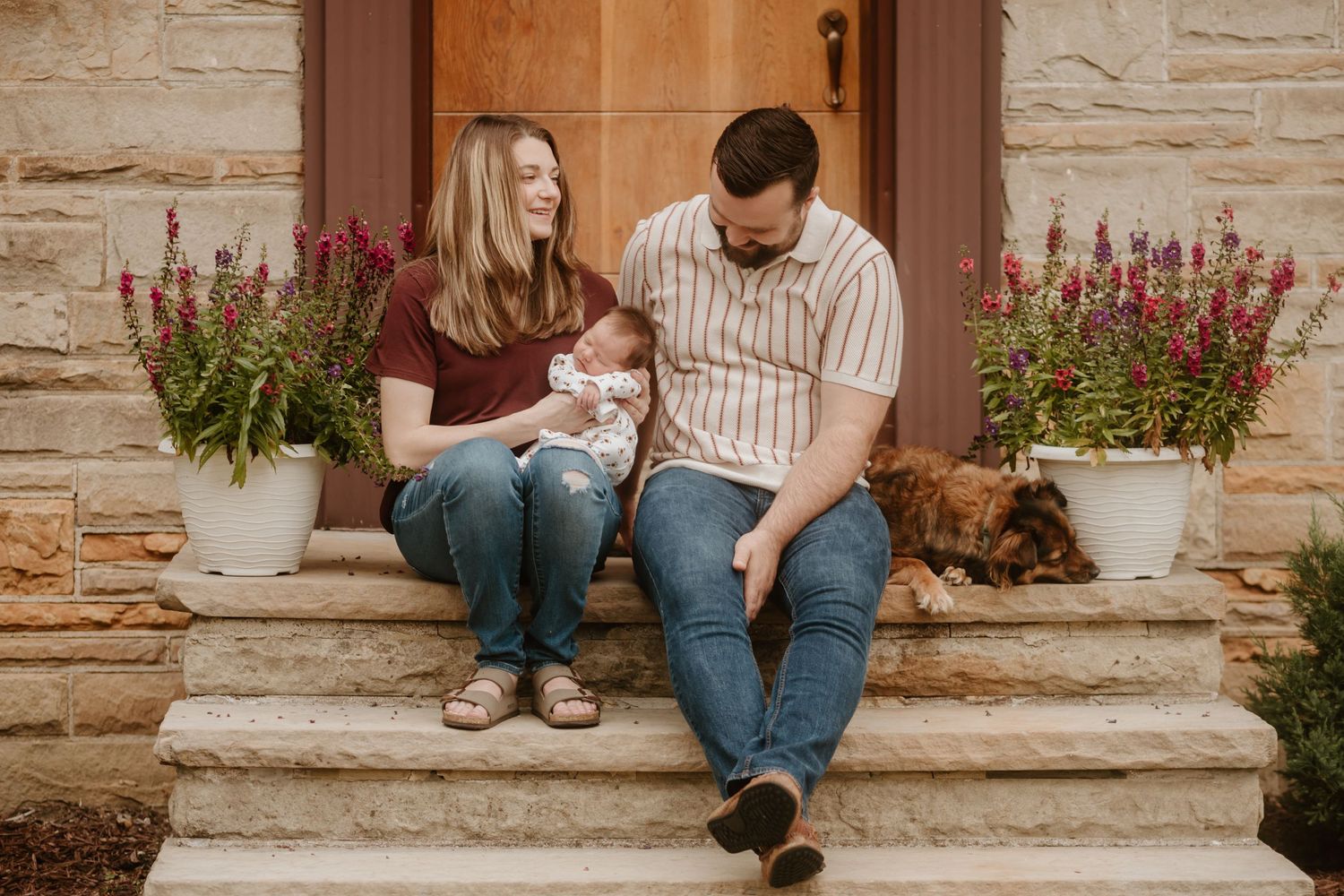 Family sitting on stone steps with potted flowers and dog by wooden door.
