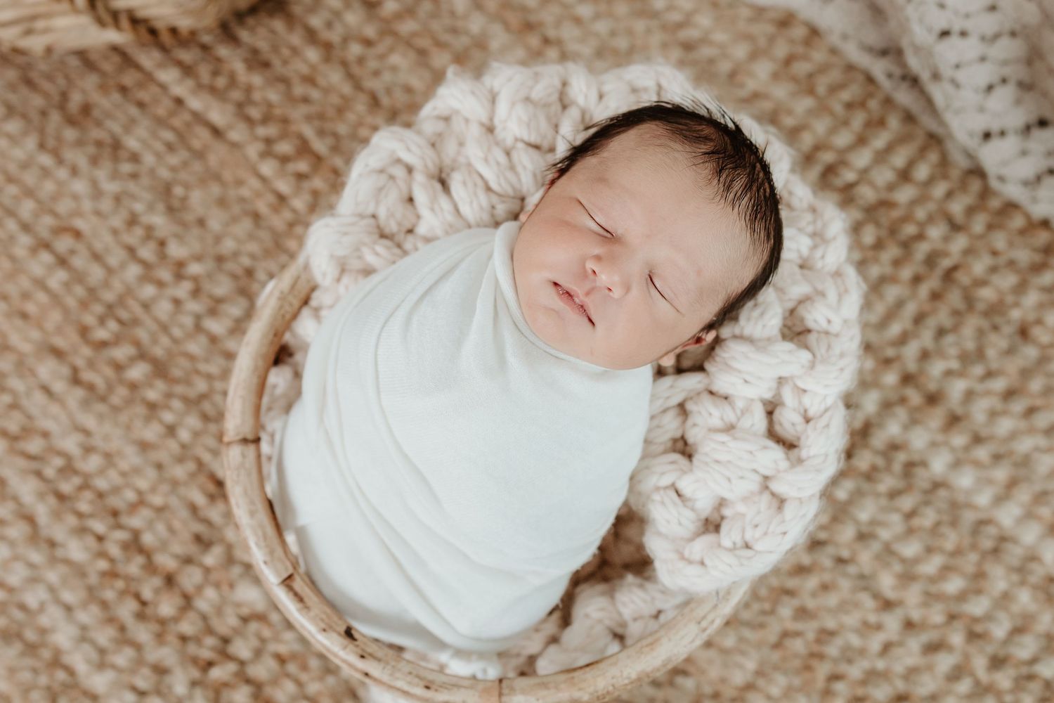 Sleeping newborn baby swaddled in white wrap lying in a cozy cream-colored knit basket.