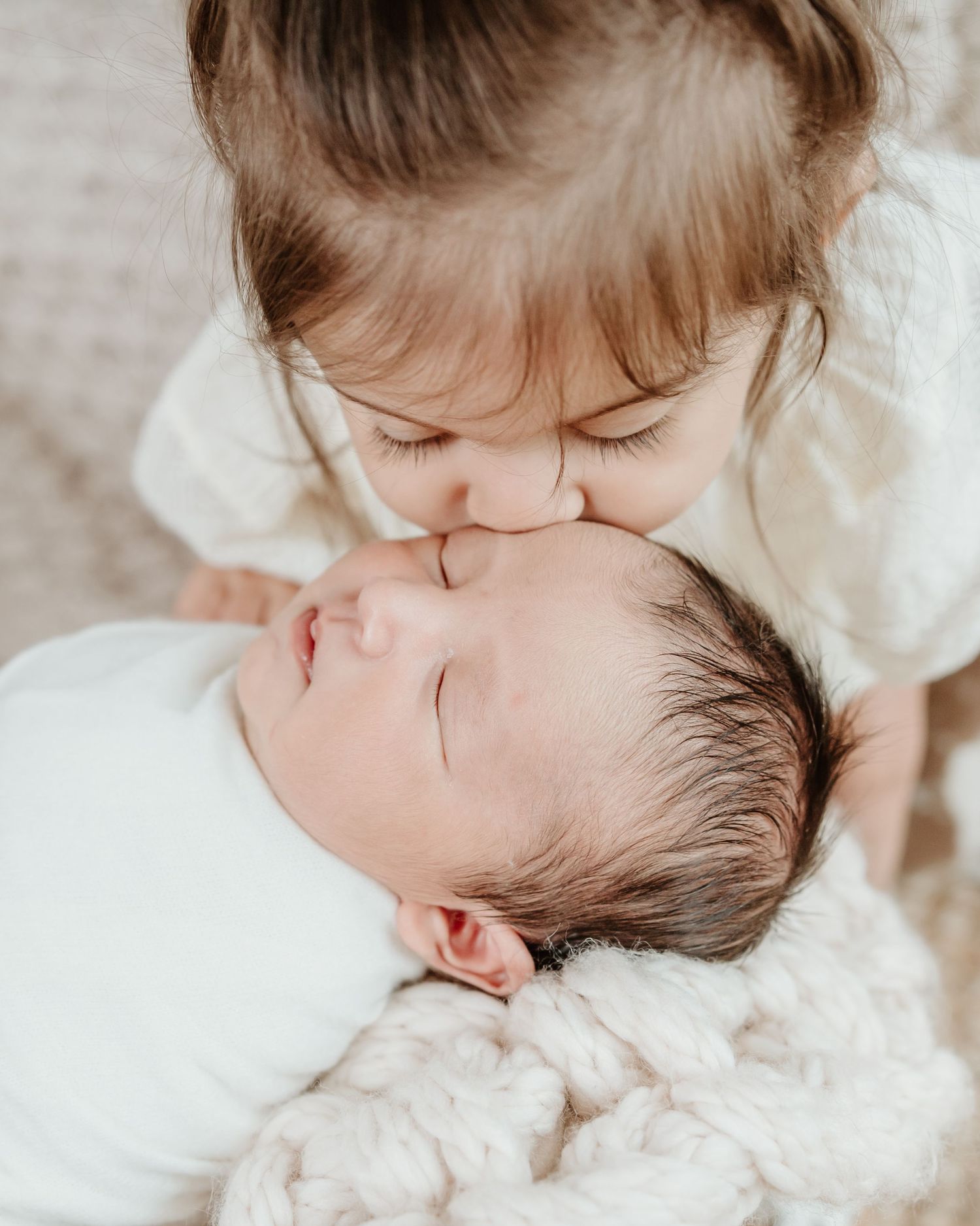 Sweet moment between siblings as older child kisses sleeping newborn on soft blanket.