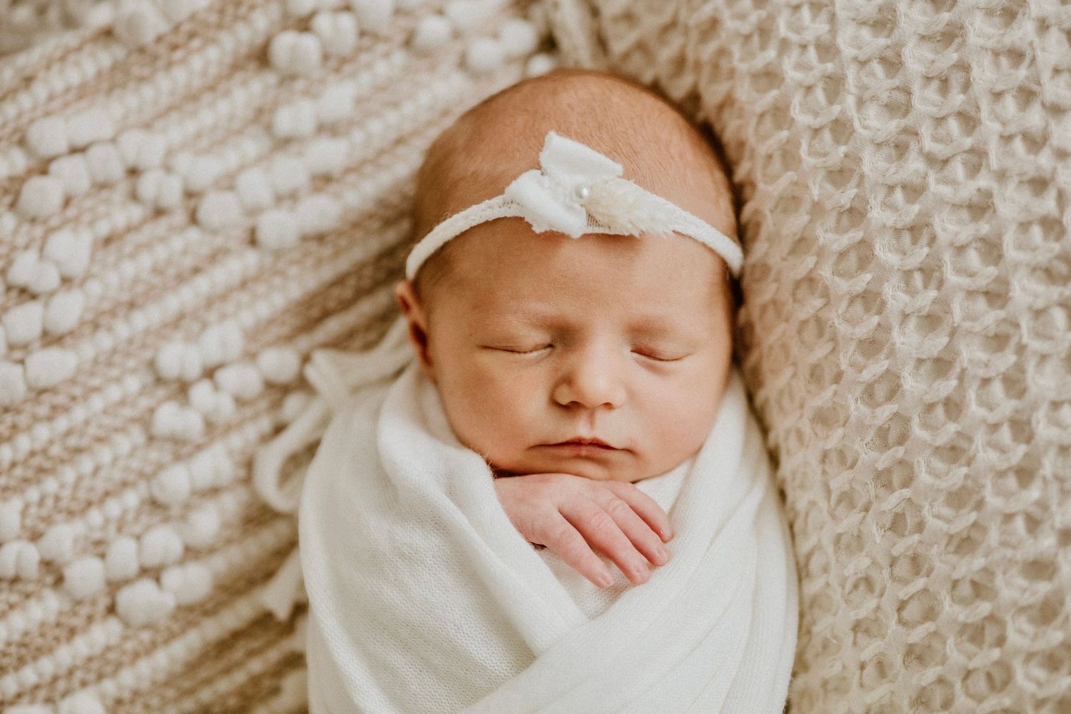 Sweet newborn baby swaddled in white with bow headband resting on cream knitted blanket.