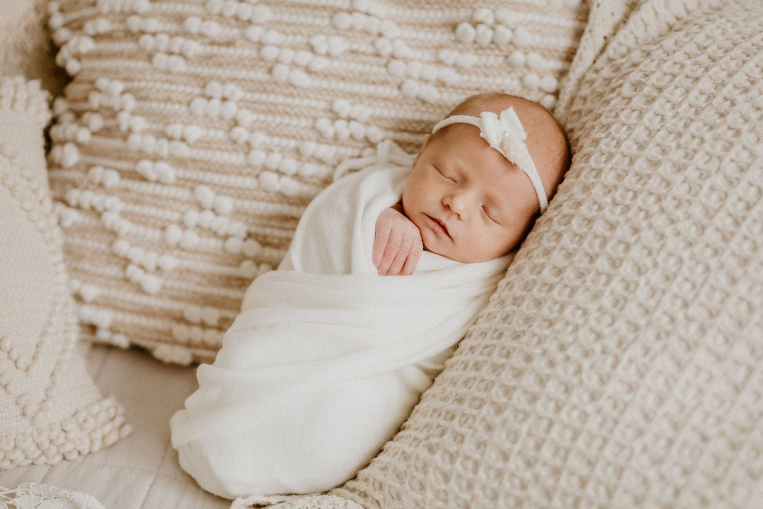 Peaceful sleeping newborn swaddled in white wrap on textured knit blanket with delicate headband bow.